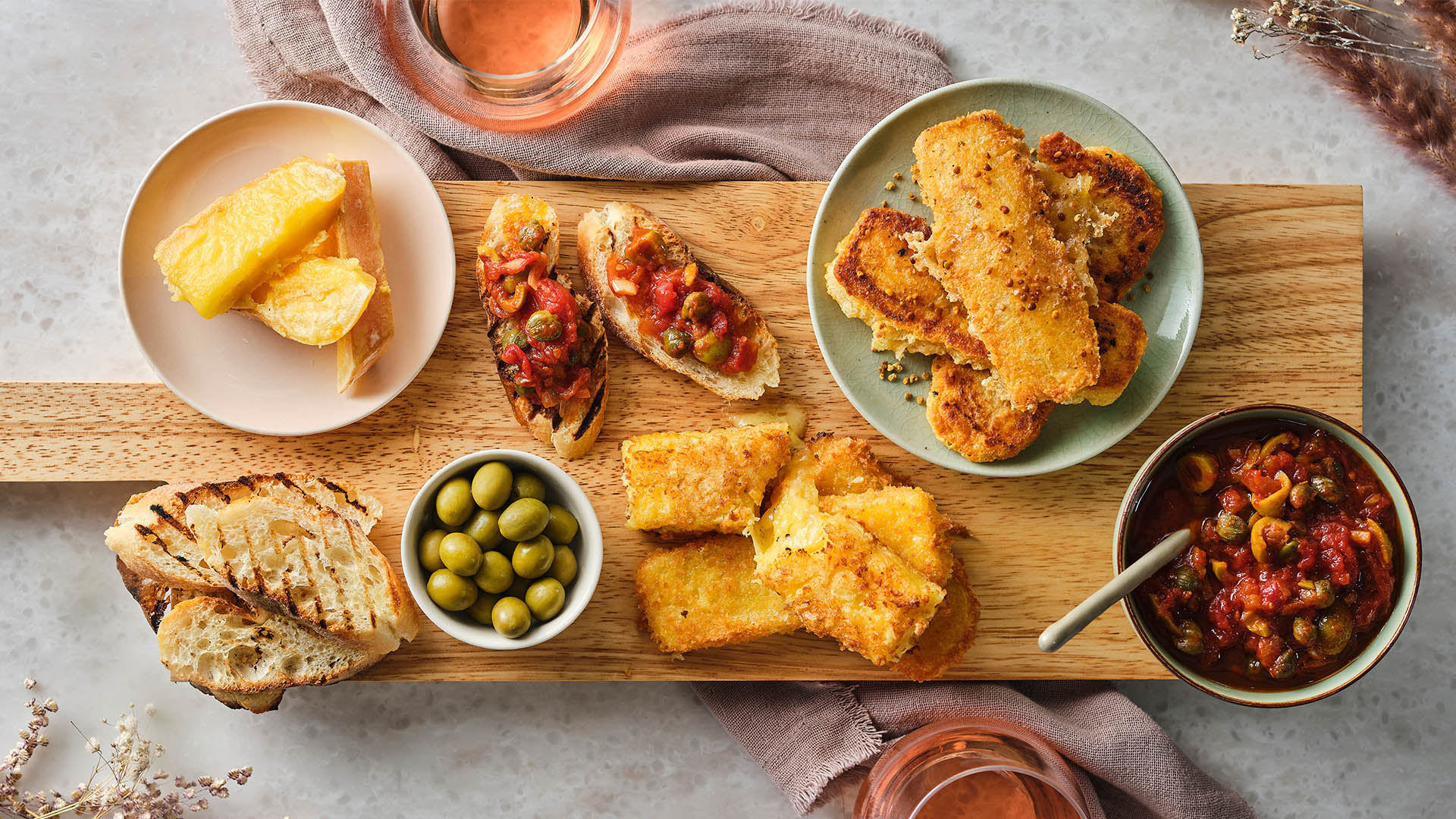 Overhead shot of a wooden serving board with pan fried cheeses, bread, bowl of olives, tapenade.