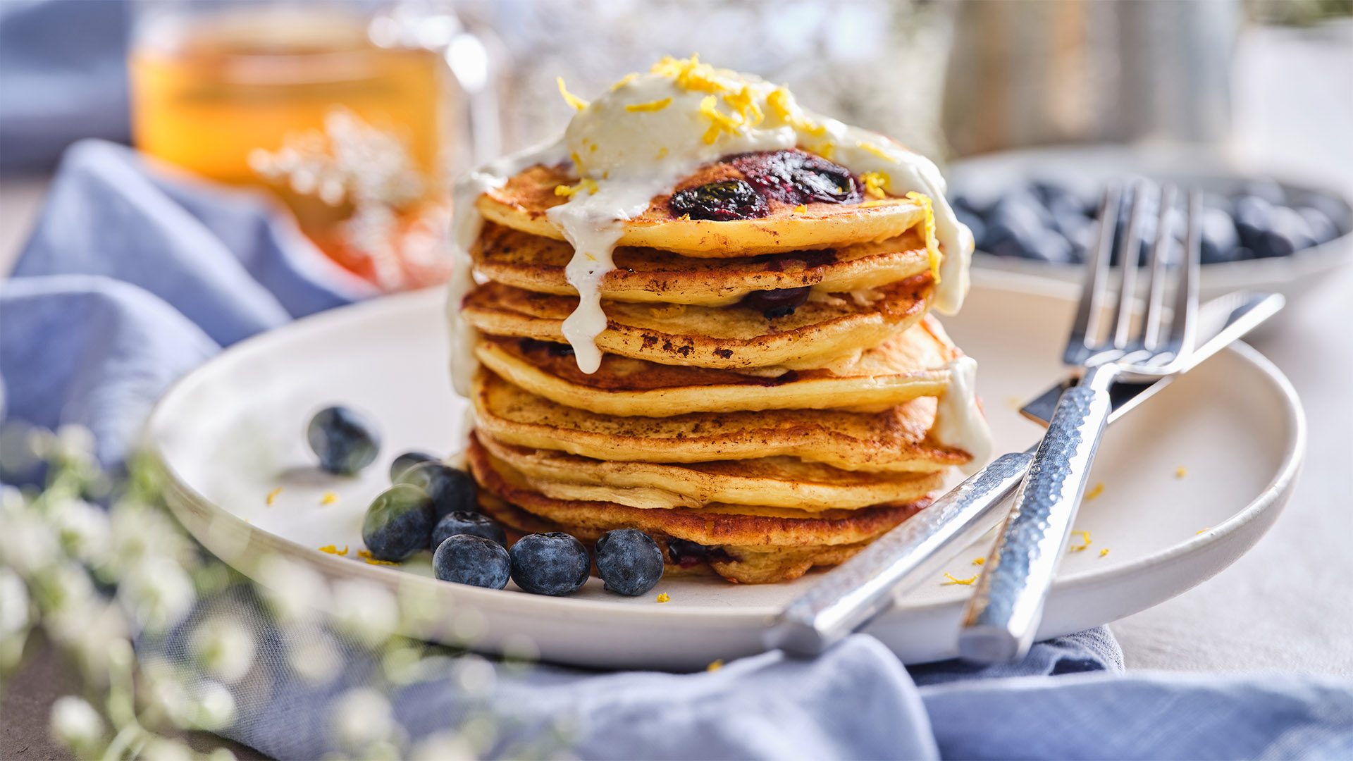 Stack of pancakes with ricotta and lemon blueberry drizzle on top on a white round plate next to a silver fork and knife.