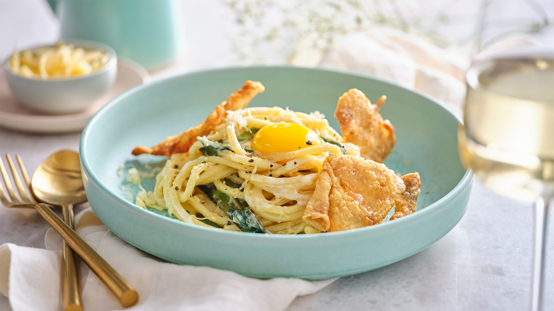 Schmaltz Carbonara with fried chicken skins, and an egg yolk on top in a blue round bowl next to gold colored fork and knife. 