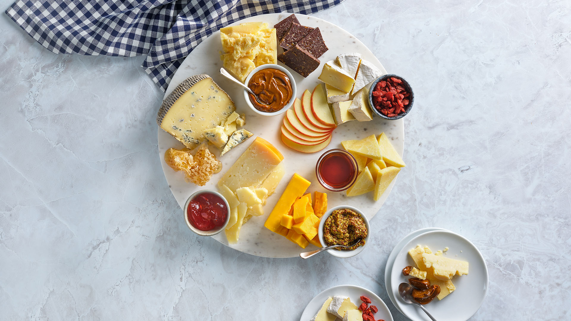 Overhead view of a round, white marble serving board with an assortment of cheese, fruit, and condiments for a charcuterie board next to a black and white checkered napkin.