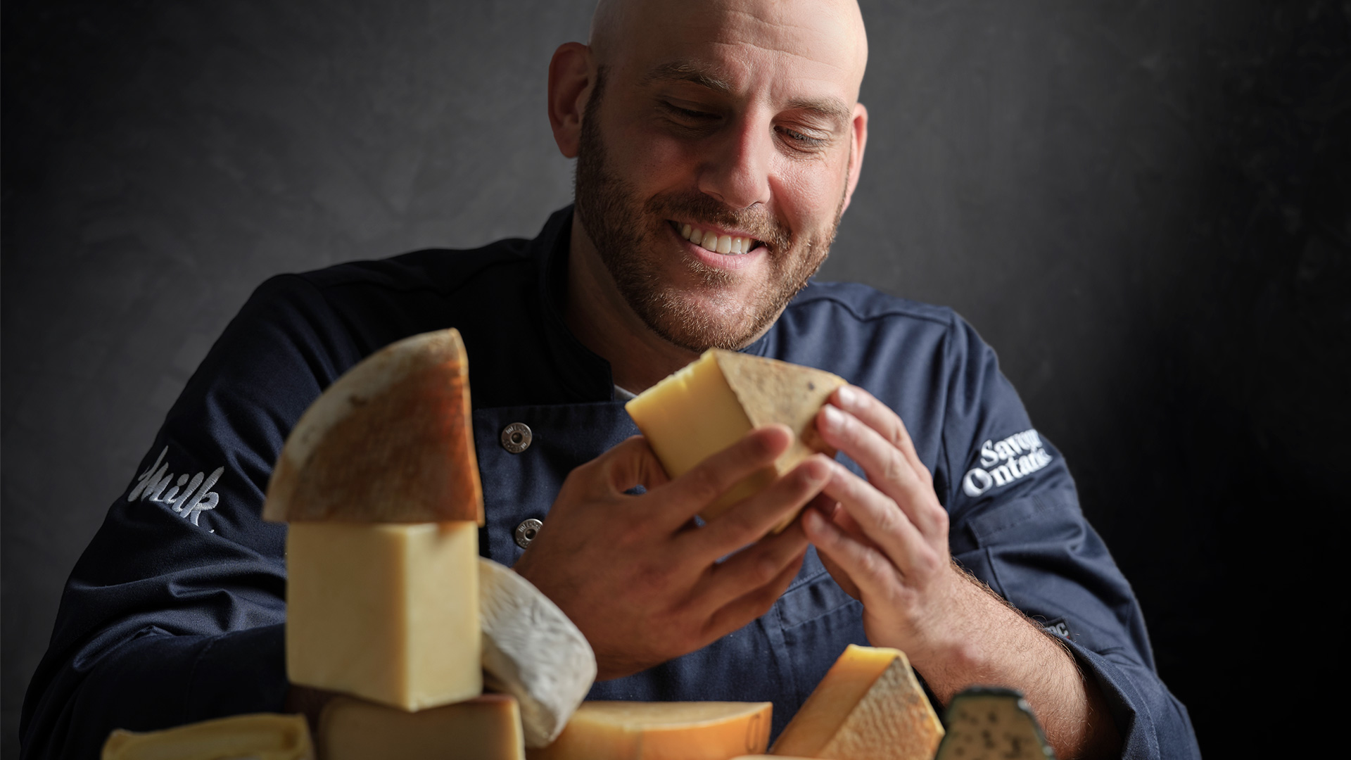 Afrim Pristine holding a wedge of cheese with multiple cheese cuts and wedges sitting on a wooden table in front of him. 