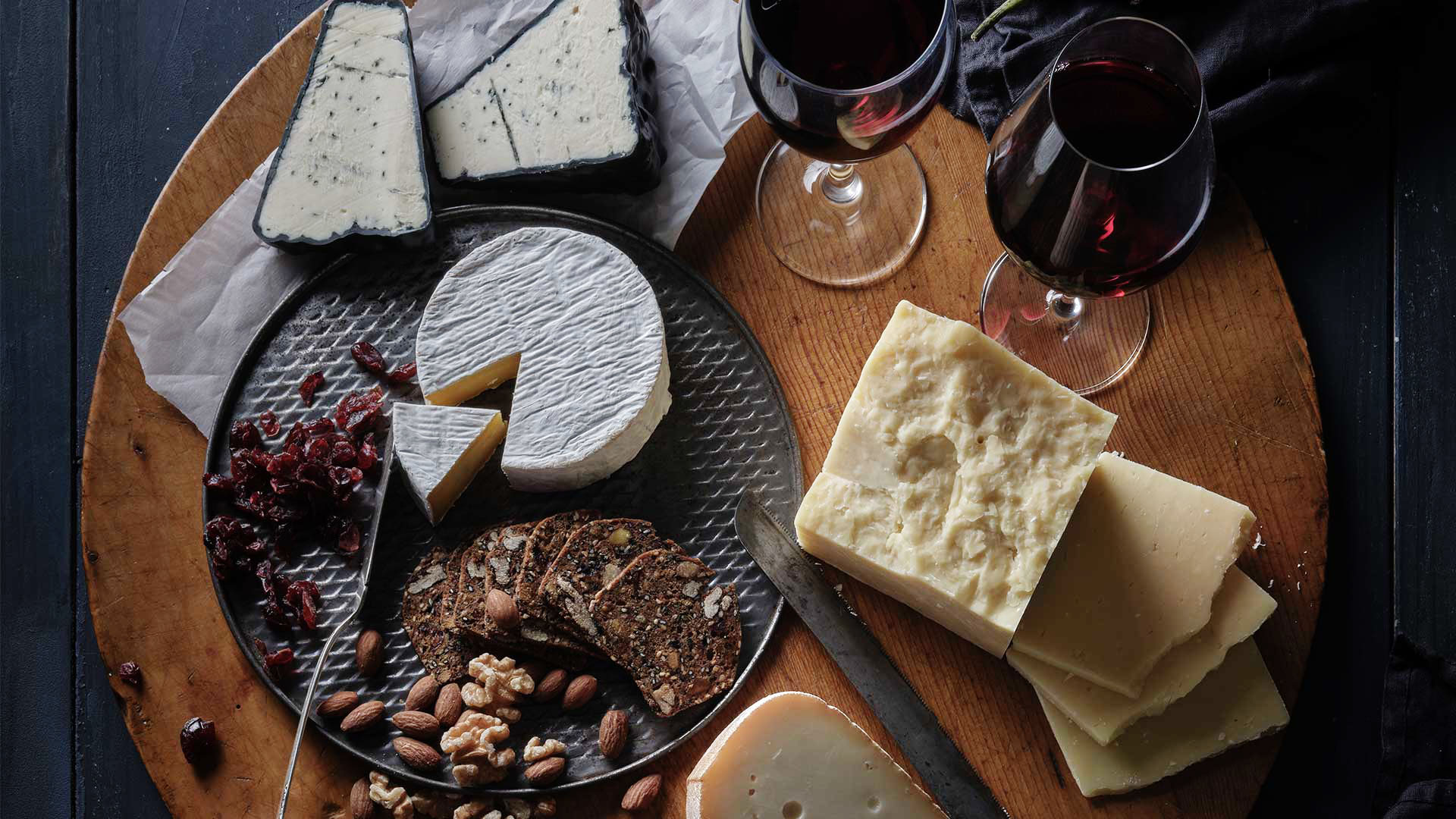 Over head shot of a round serving board with various types of artisanal cheeses and two glasses of wine.