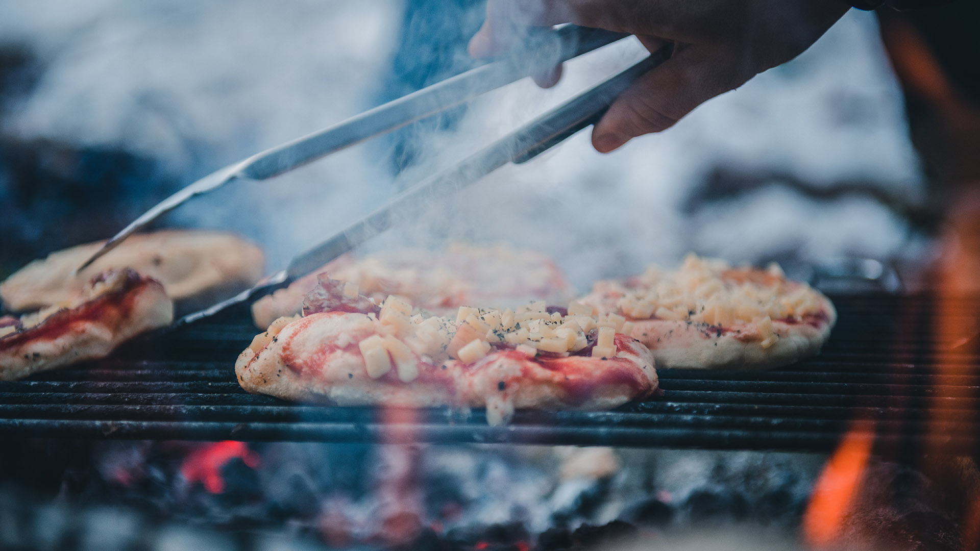 Small pizzas cooking on a grill over a campfire with a hand holding tongs grabbing the edge of one of the pizzas.