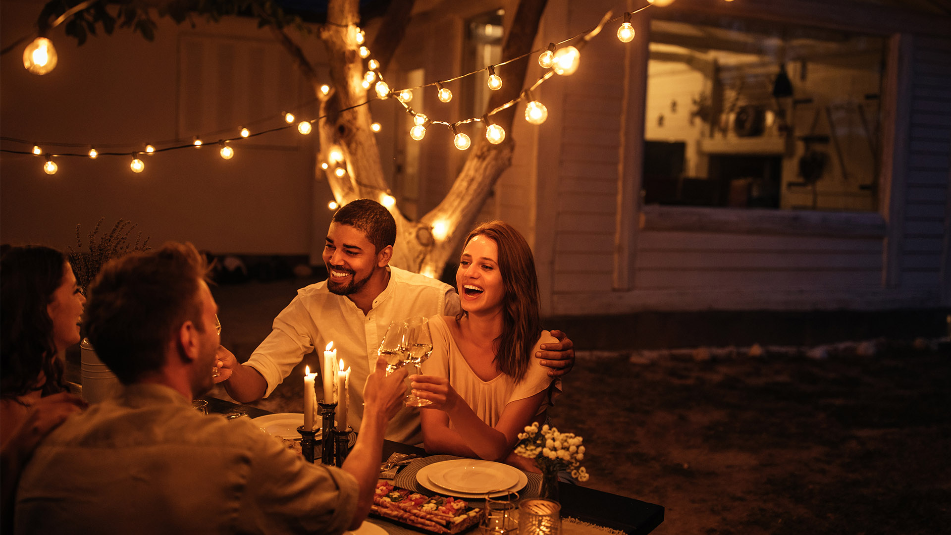Two couples holding up their glasses for a toast at a picnic table under string lights in a backyard.  