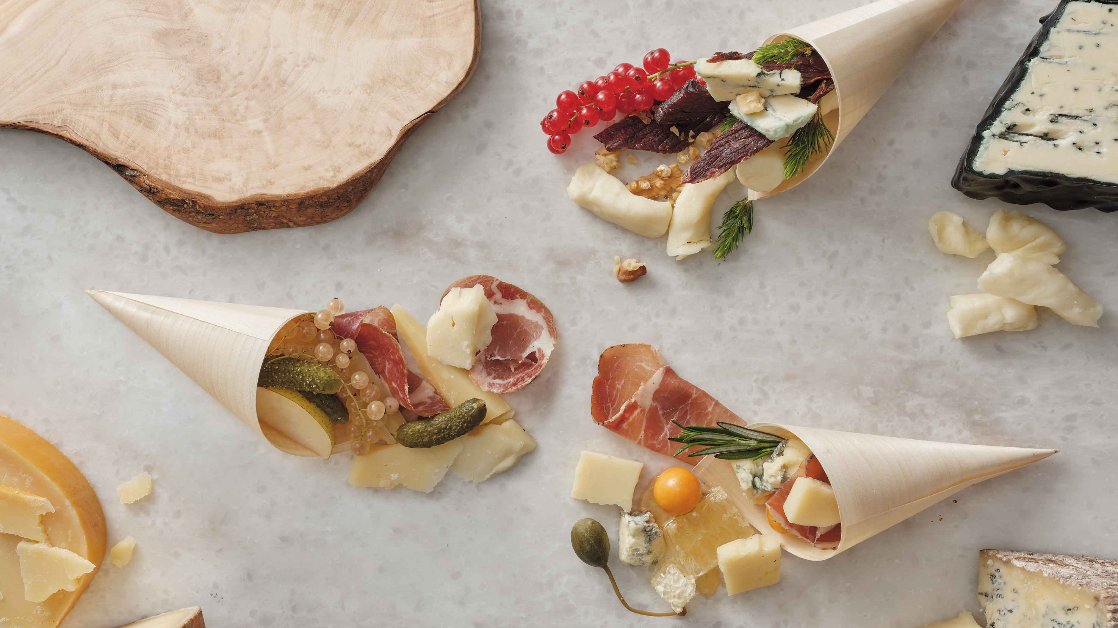 Overhead view of three bamboo cones filled with artisanal cheeses, gherkin pickles, currants, honeycomb, and cured meats.