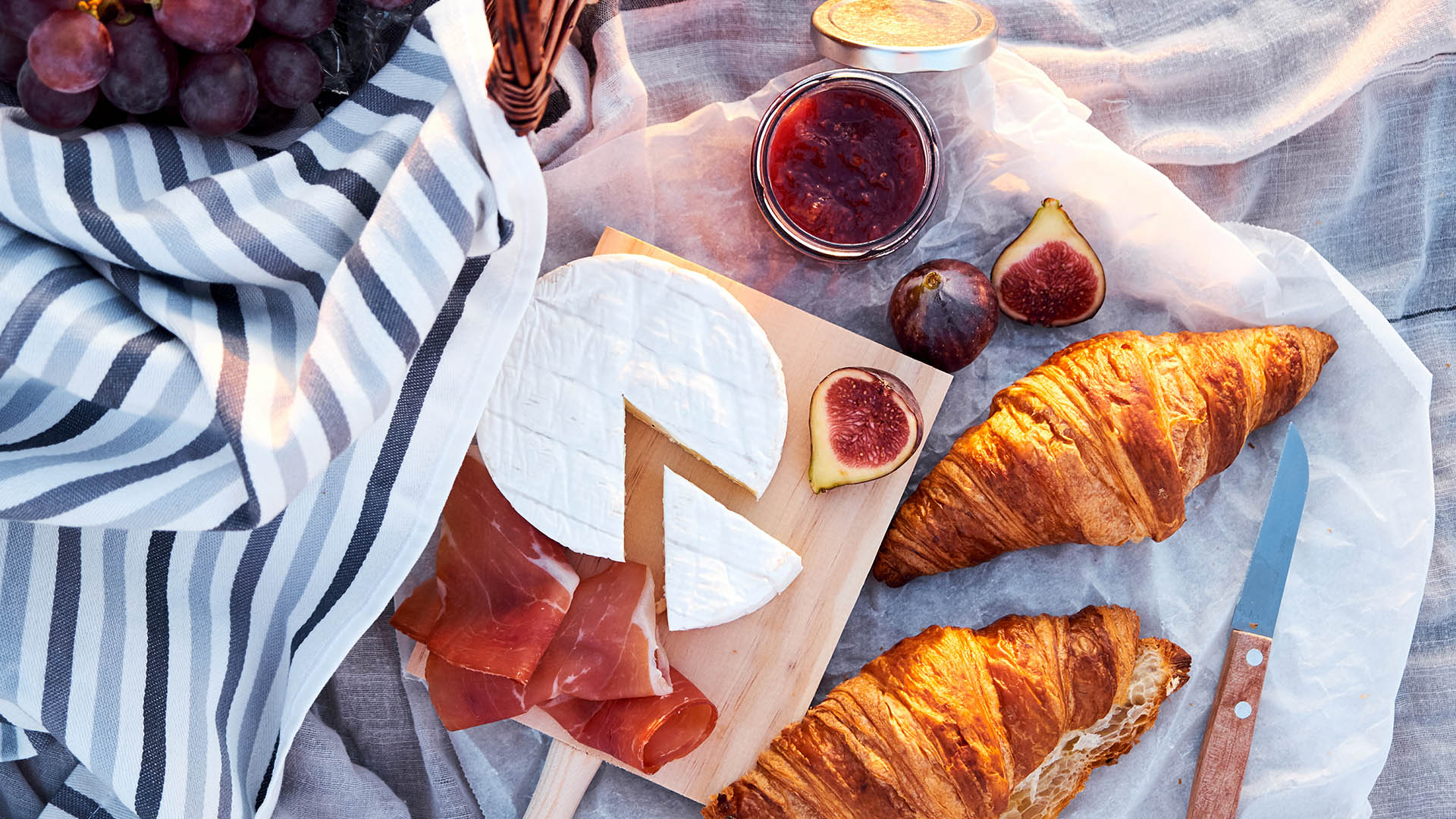 A white picnic blanket on the ground with a brown wicker picnic basket with bread, grapes and wine glasses inside. To the right there is a wheel of Brie and some croissants on a cutting board