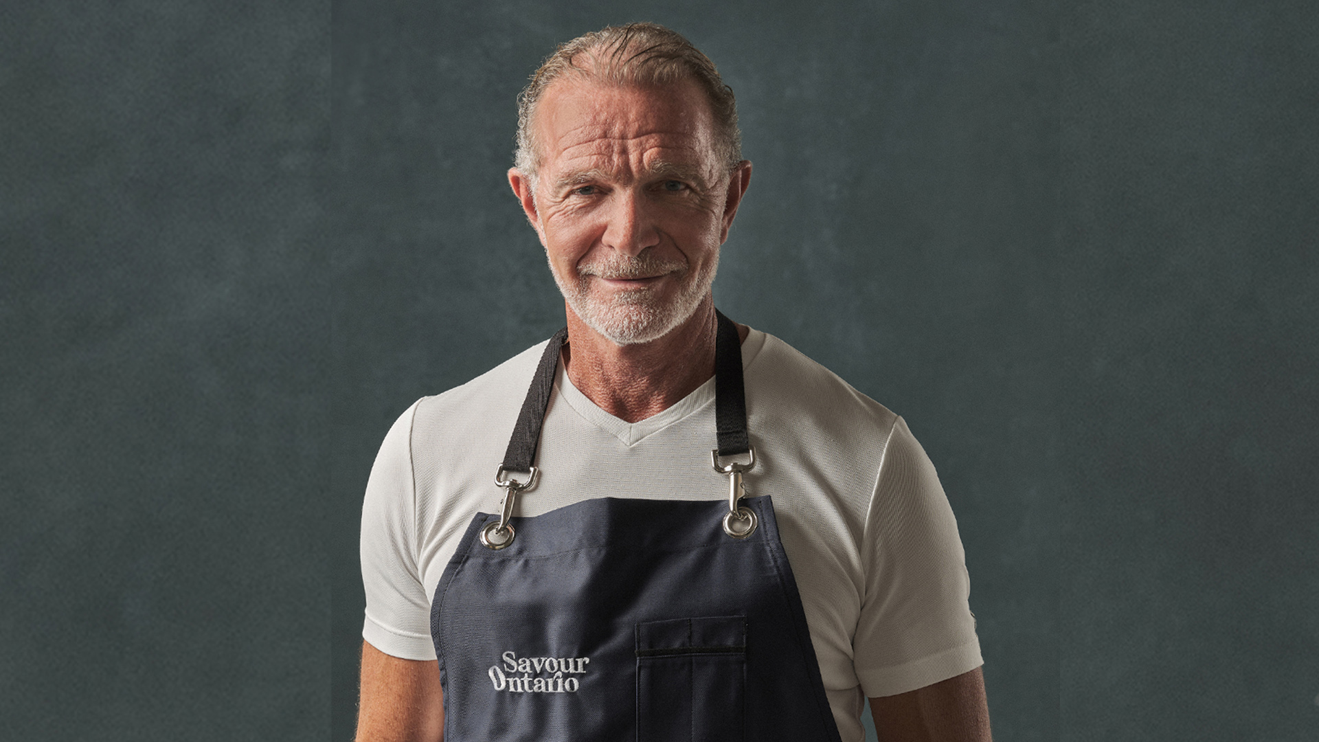A headshot of Chef Mark McEwan wearing a short sleeved white t-shirt underneath a navy Savour Ontario apron.