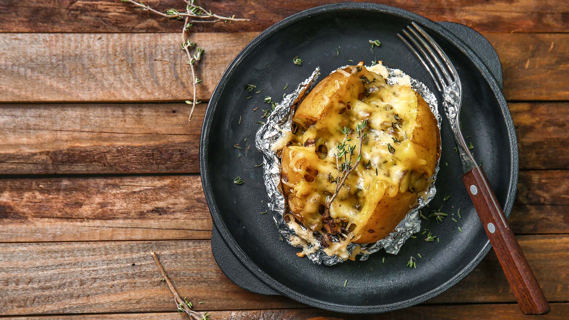 Overhead view of cheesy baked potato in tin foil next to a fork on a round black plate on a wooden surface. 