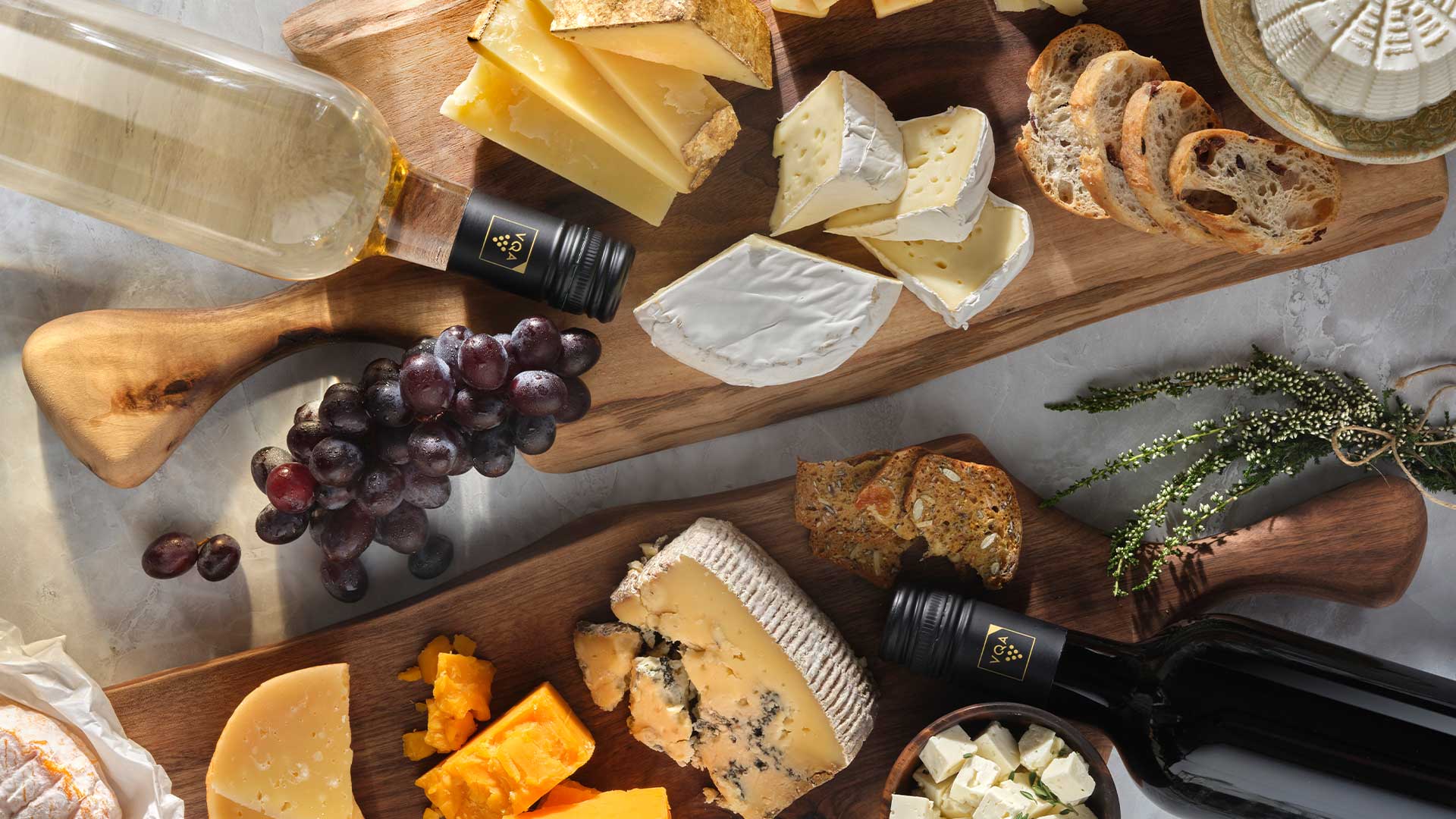 Overhead view of two charcuterie boards with different varieties of artisanal cheeses and two VQA wine bottles, one is red wine the other is white wine. 