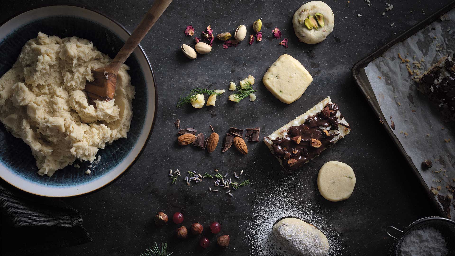 Overhead view of a large bowl filled with shortbread cookie dough with a wooden spoon sticking out of it, and 5 lines of various cookie dough ingredients such as pistachios, rose petals, dill, almonds, chocolate chunks, lavender, and hazelnuts. 