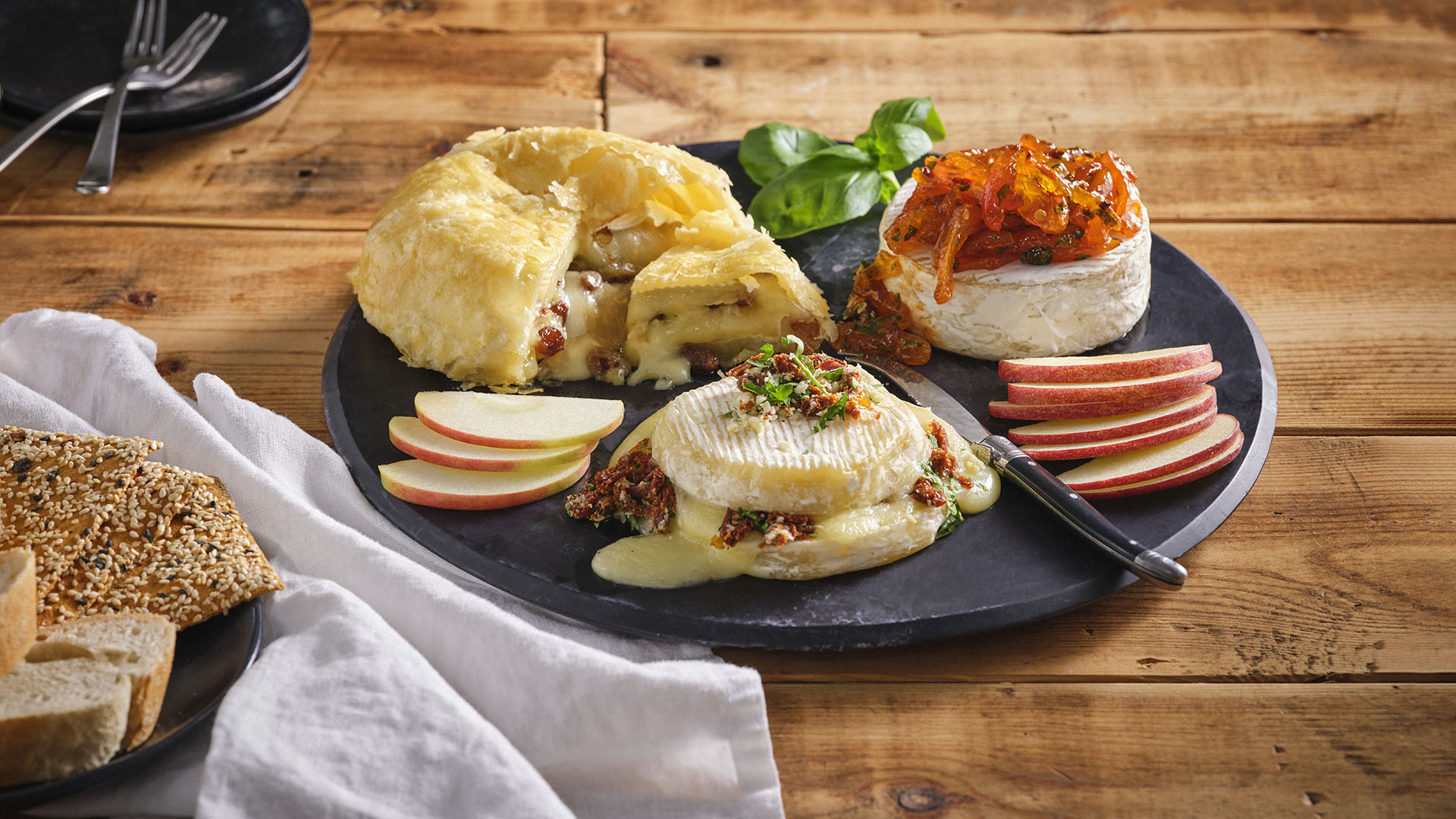 Round, black serving board with three baked bries, one with wrapped in pastry dough, one with dried apricot on top, and the other stuffed with sun-dried tomato, asiago and parsley.