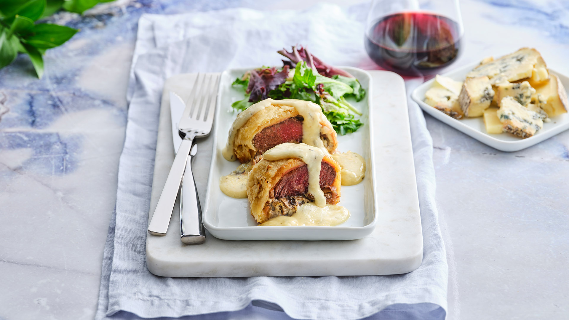 Beef and Blue Cheese Wellington on a rectangular, white plate next to a plate of crumbled blue cheese next to a small wedge, and a glass of red wine. 