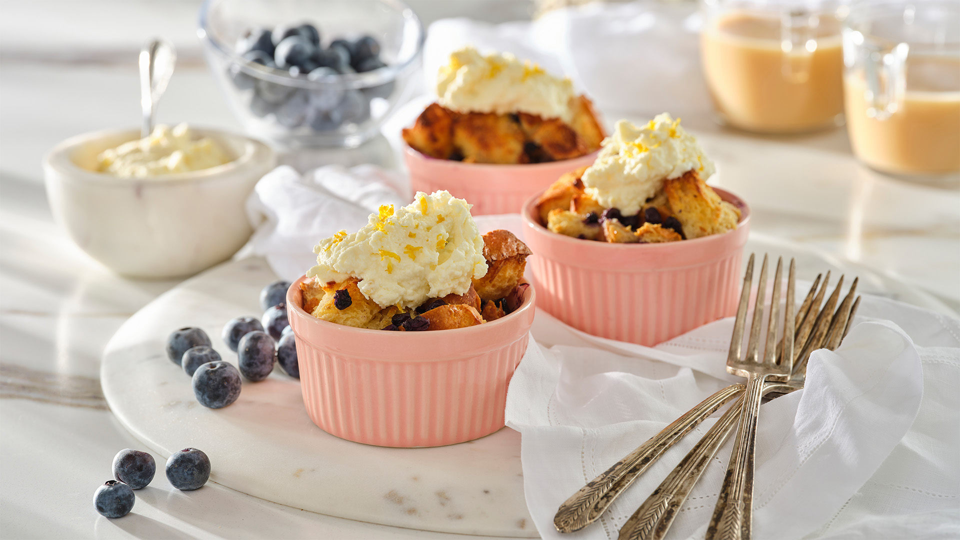 Three small pink bowls of blueberry french toast bake with a dollop of lemon mascarpone on top next to some blueberries, forks, and a small white bowl containing mascarpone with a spoon in it.