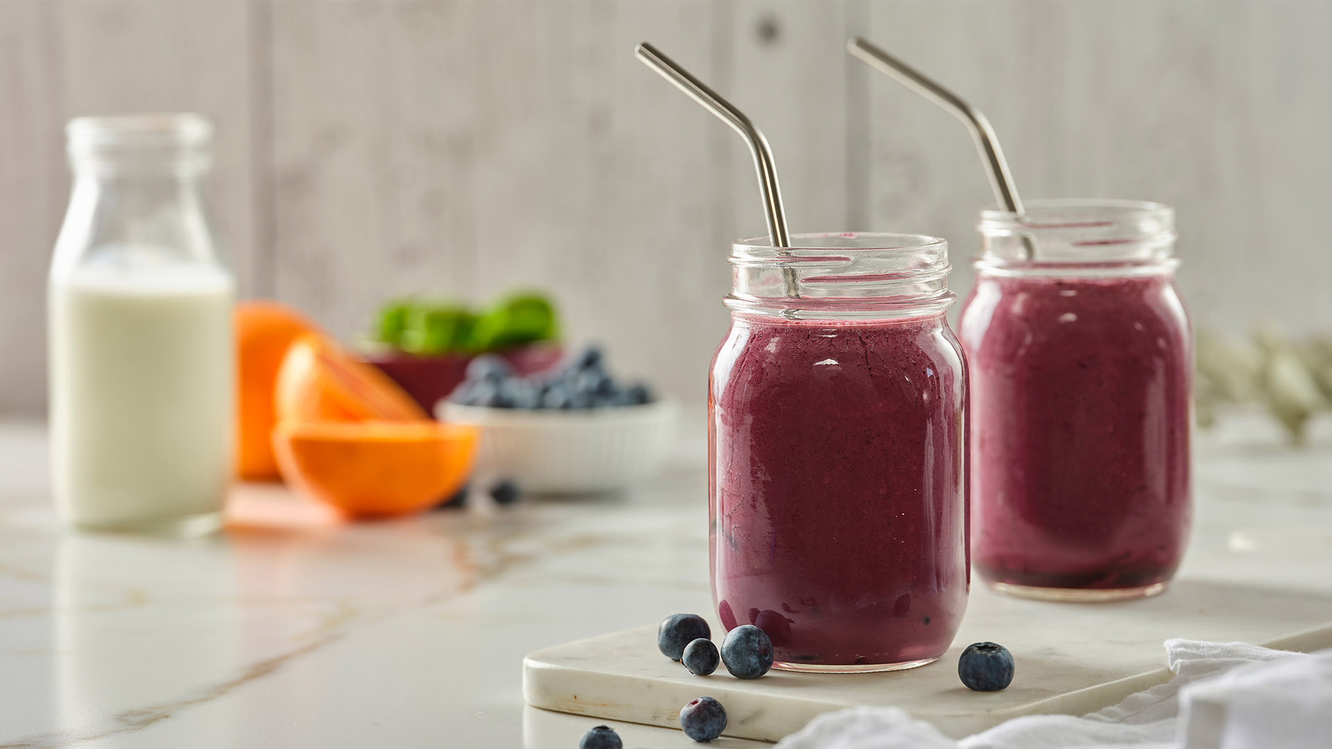 Side shot of two jars with metal straws containing blueberry kefir smoothie with pitcher of milk and small bowls with smoothie ingredients in them in the background.