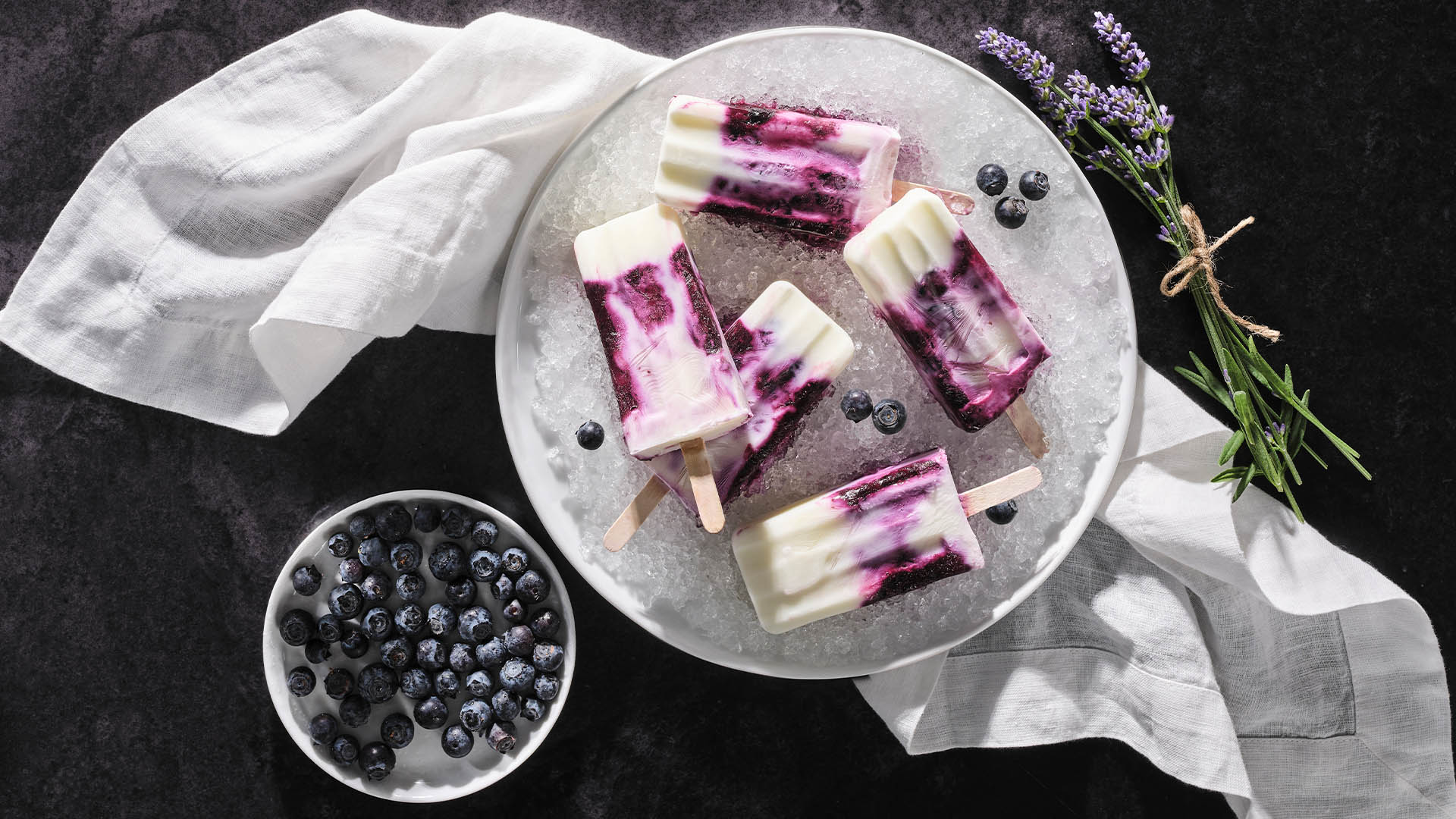 A round white plate with blueberry lavender yogurt popsicles on a bed of crushed ice, with blueberries strewn around the popsicles. A small white plate with fresh blueberries and a bundle of fresh lavender. 