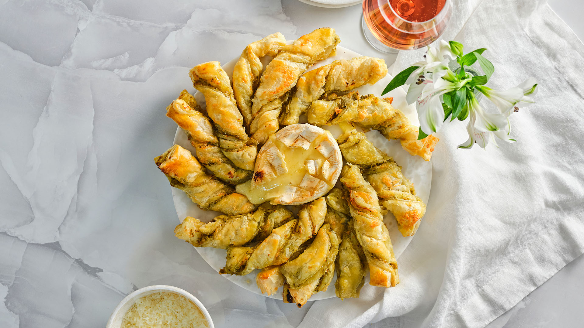 Overhead view of Pesto pastry twists surrounding a baked wheel of camembert on a round marble serving board.
