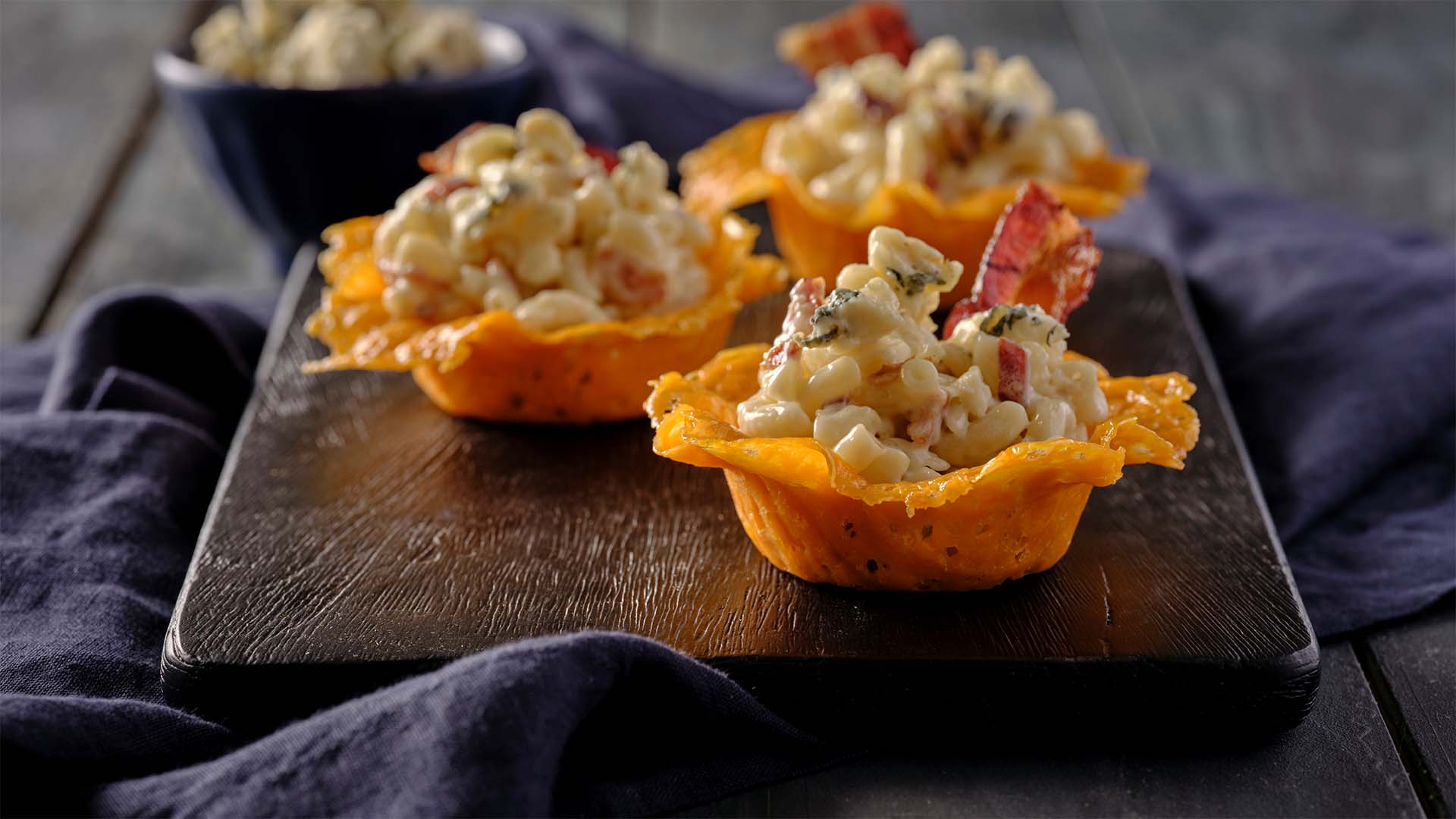Three Mini Gourmet Mac n' Cheese cups sitting on a black serving board filled with macaroni and cheese topped with blue cheese and bacon.