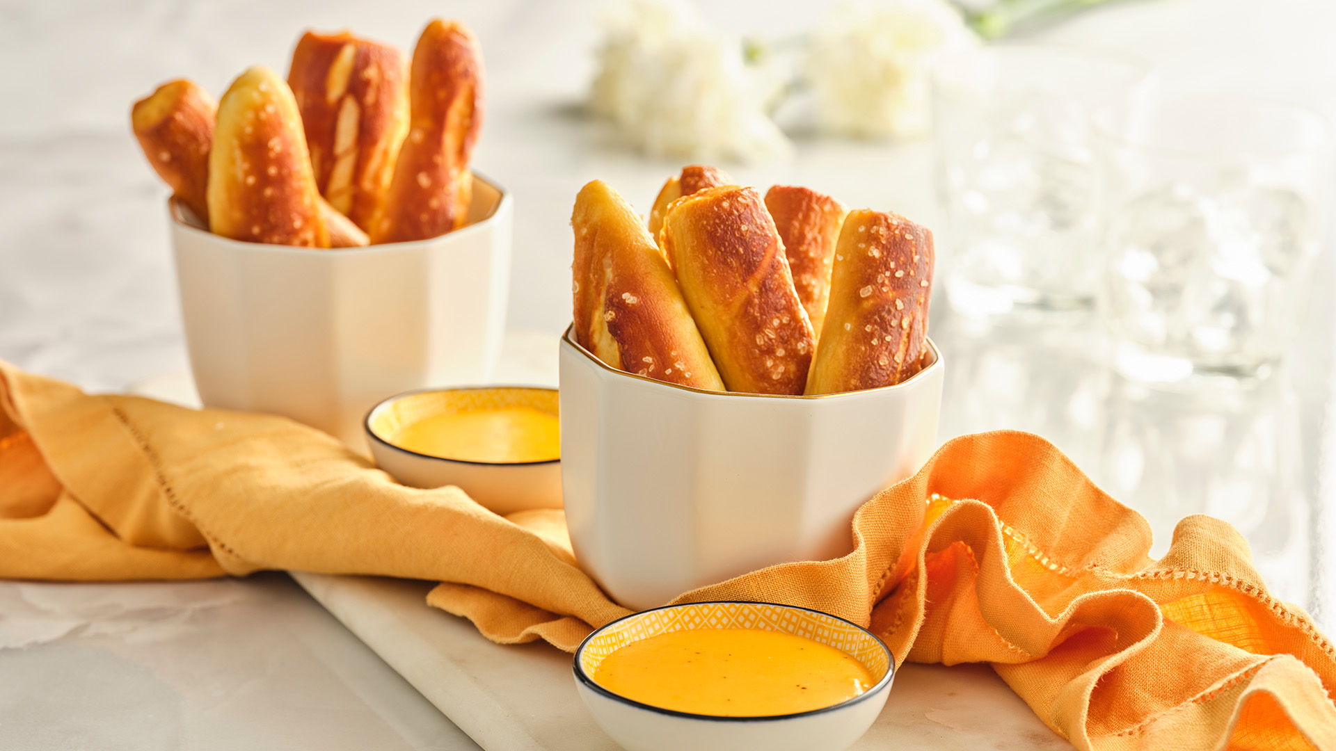 Two white bowls with homemade pretzel sticks next to two small white bowls containing cheese dip and an orange napkin.