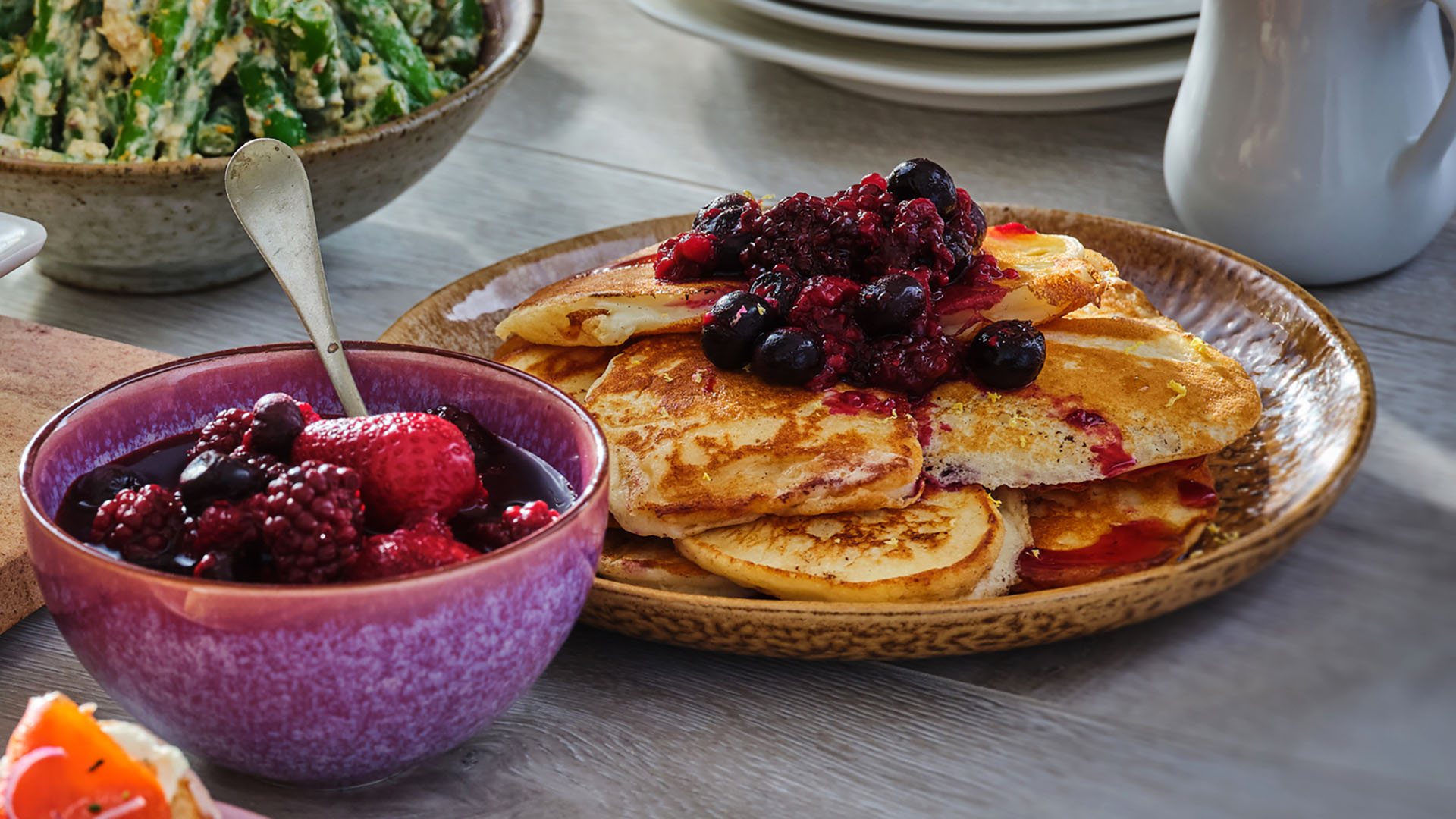 Lemon ricotta pancakes topped with fruit on a round purple dish with a bowl of fruit to the left of the plate.