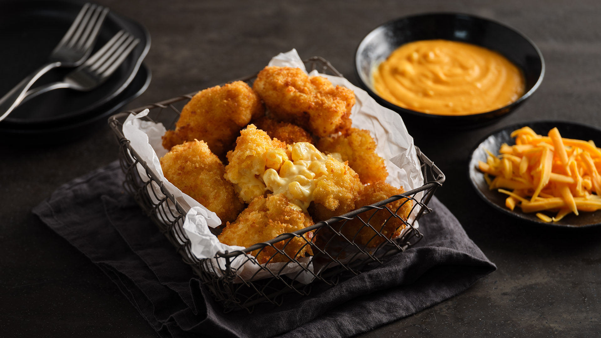 Macaroni & Cheese Croquettes on top of parchment paper in a metal, wire basket next to a round dark bowl of shredded cheese and another round bowl with cheese dip.