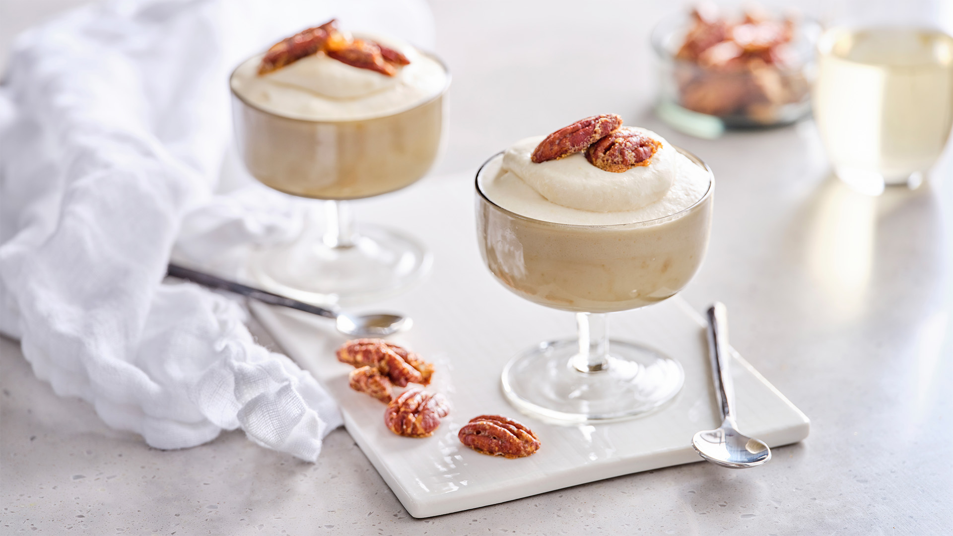 Two Maple mousse filled glasses on white serving board next to some pecan pieces, small silver spoons and a white napkin.