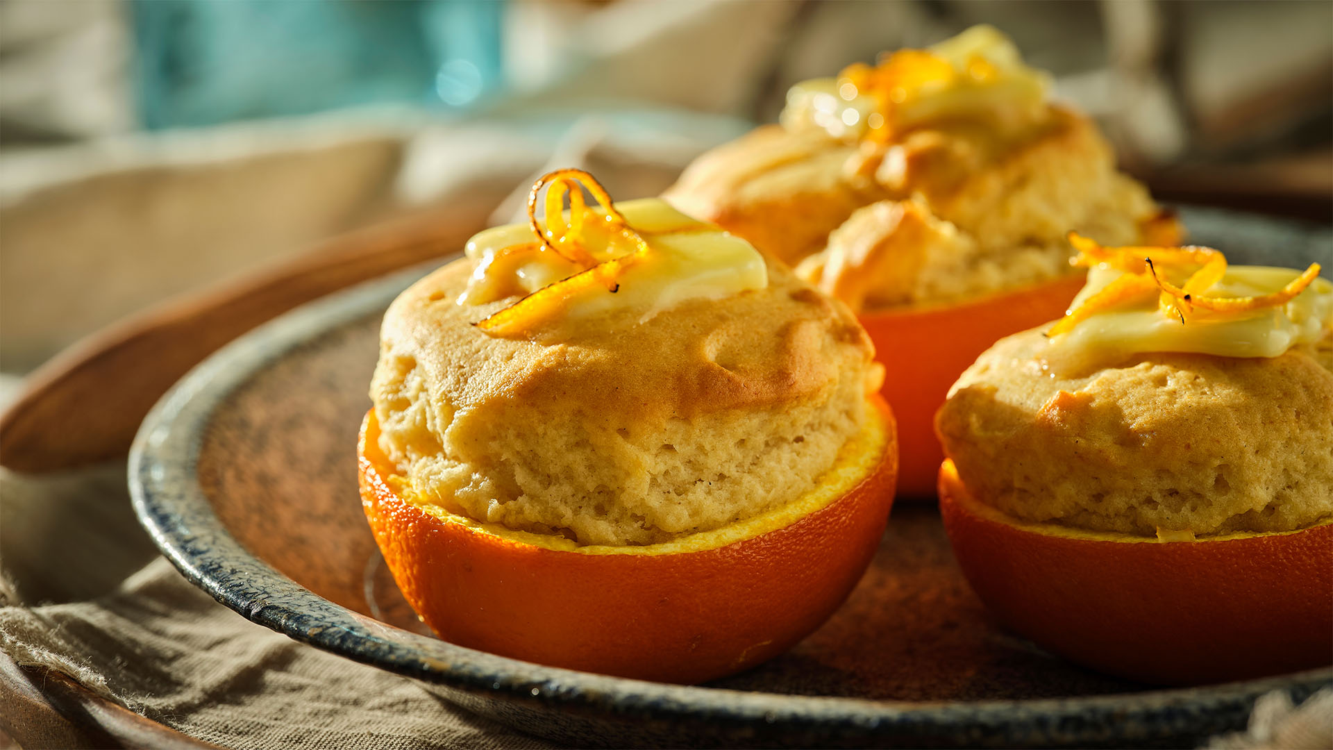 Three orange baked spelt muffins topped with butter and orange peel twist on a round enamel plate. 