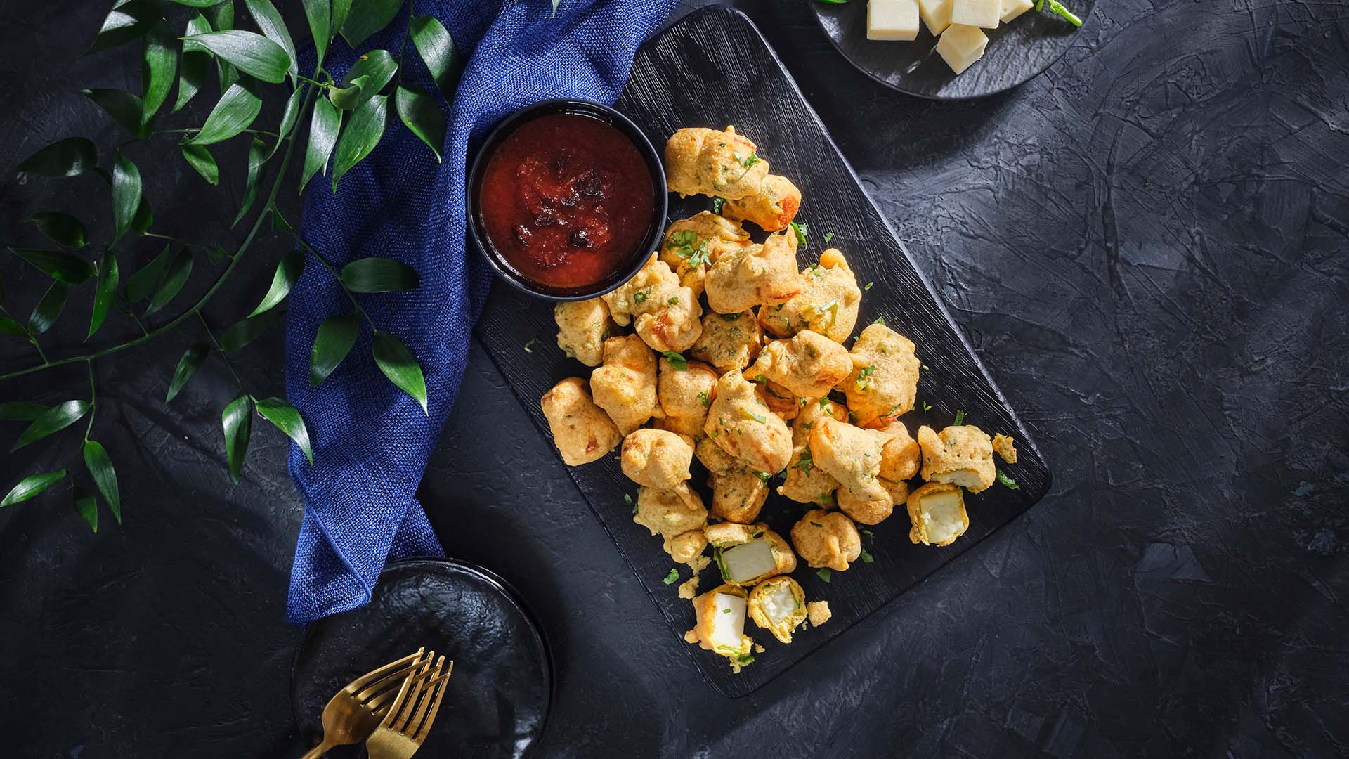 Overhead view of Paneer Pakora on a black, rectangular serving board, one broken open exposing paneer inside next to a small bowl of tomato chutney,