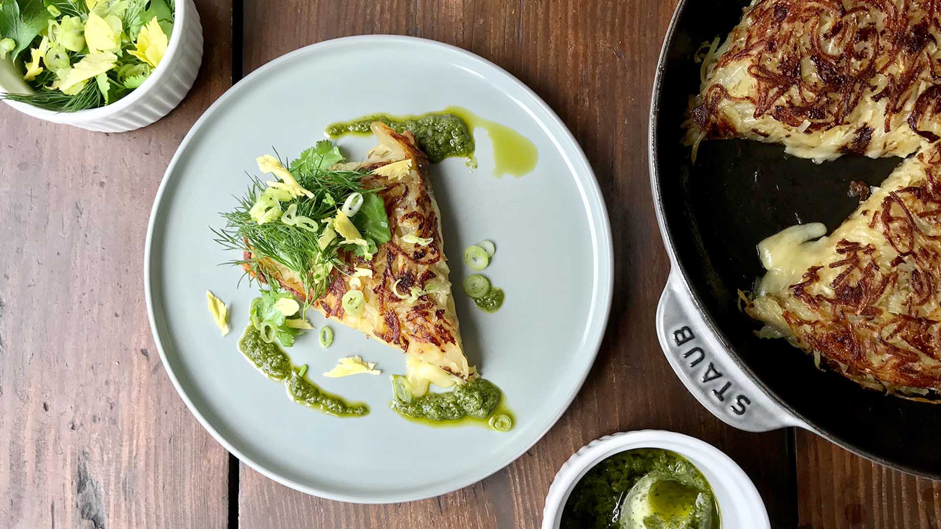 Overhead shot of potato rosti with green drizzle and green onion garnish on round white plate on wooden surface next to pan of rosti and two small white bowls.