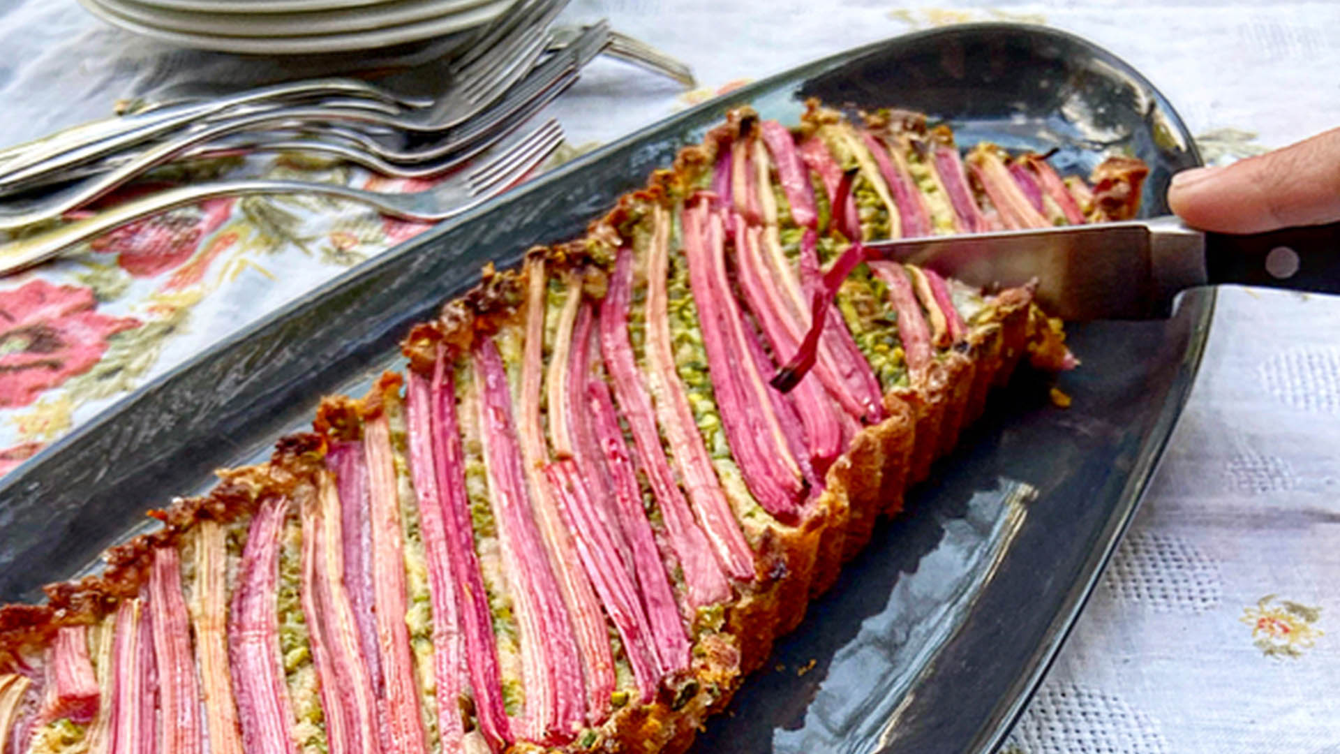 Rhubarb pistachio cake on black long oval shaped serving plate with knife cutting into it.