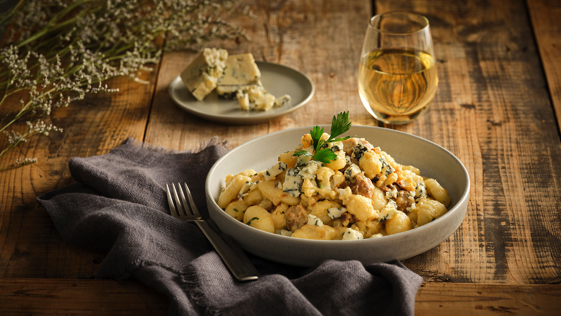 Sausage Gnocchi with Ontario Feta & Roasted Cauliflower in a shallow, round, white bowl next to a grey napkin with a fork on top, in behind is a glass of wine and a small round plate with crumbled blue cheese.