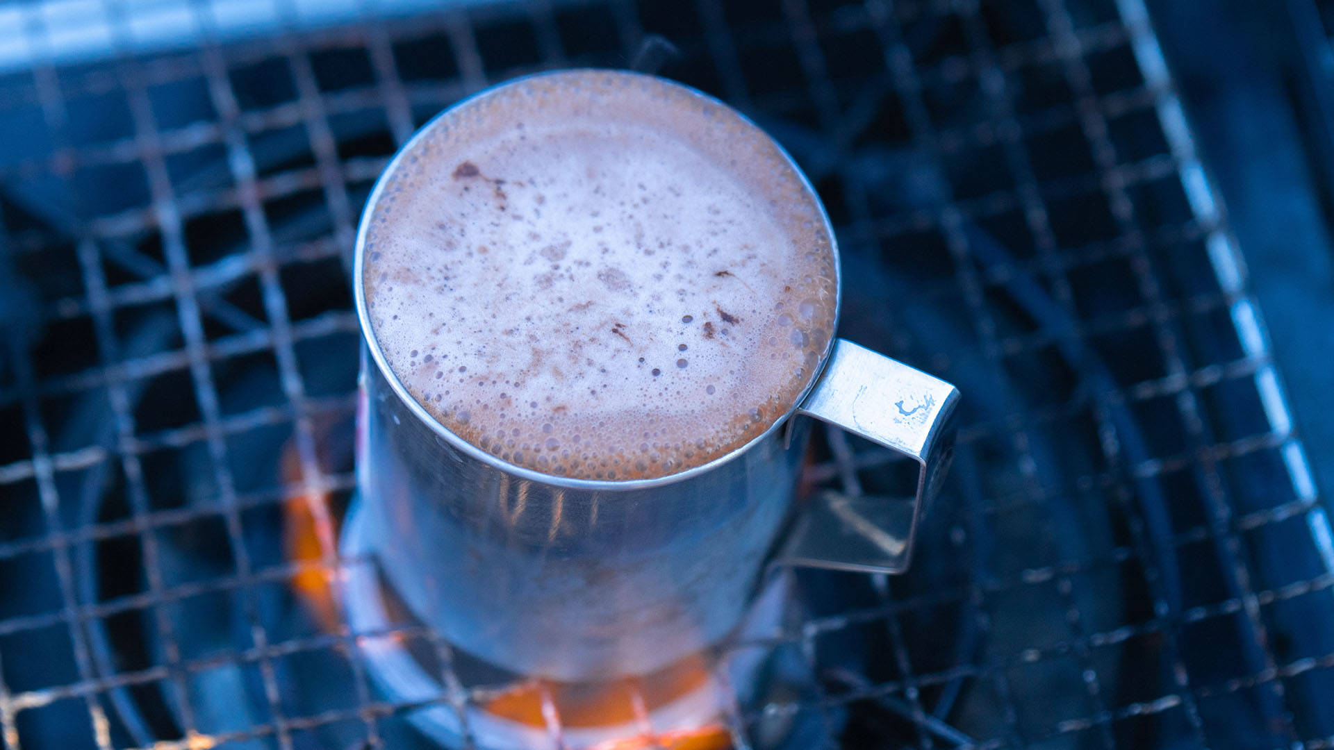 A metal mug on a camp grill full of hot cocoa.