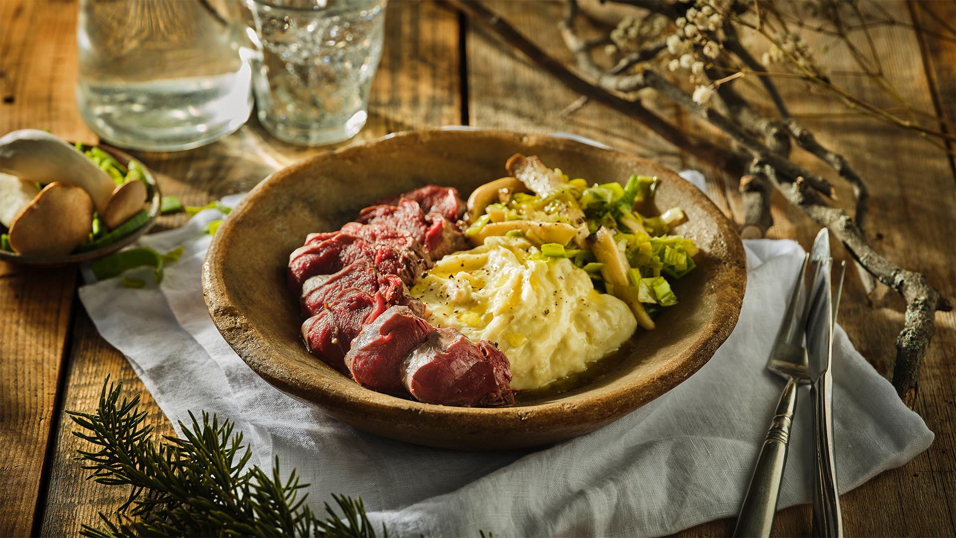 Round wooden bowl with mash, venison slices, and veg on a white napkin next to a knife and fork.