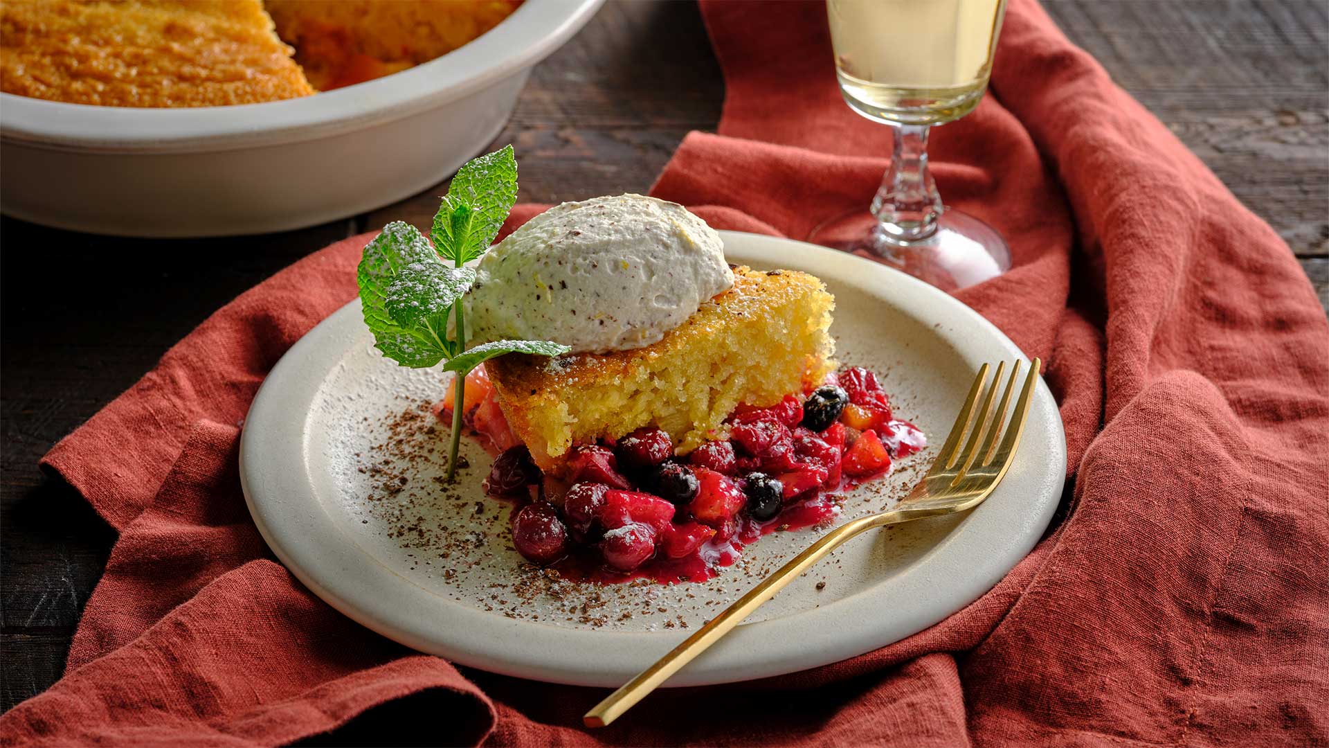A slice of sponge cake on a bed of fruit with a dollop of ricotta on top with a sprig of mint next to it, the slice of cake is on a round white plate that is set on top of a red napkin. 