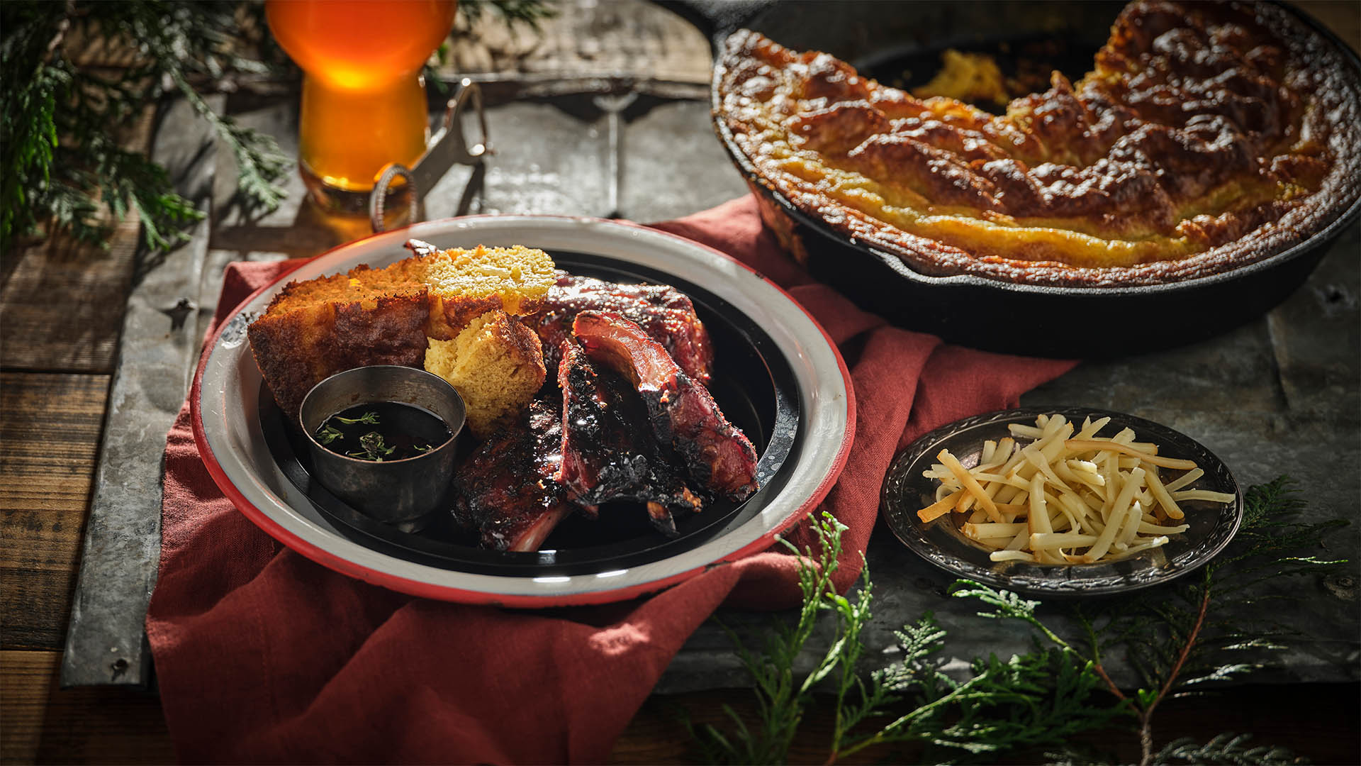 Smoked cheddar cornbread and ribs with dip on a round plate with a red rim on top of a red napkin next to a small round black plate with shredded cheese next to a cast iron pan with cornbread. 