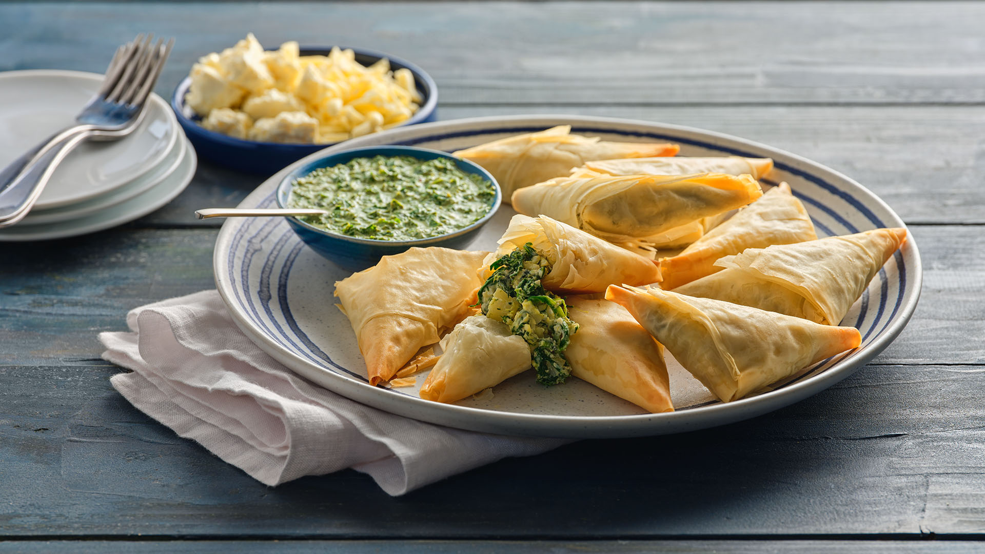 Samosas on a round plate with blue stripes next to a stack of white plates with forks on top, and a small round bowl filled with crumbled cheese.