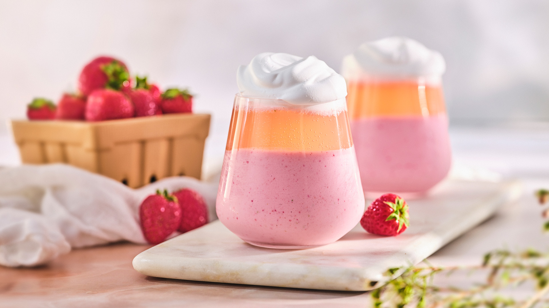 Two stemless wine glasses containing strawberry milkshake topped with Rosé and a dollop of whipped cream next to a small brown fruit pint full of strawberries.