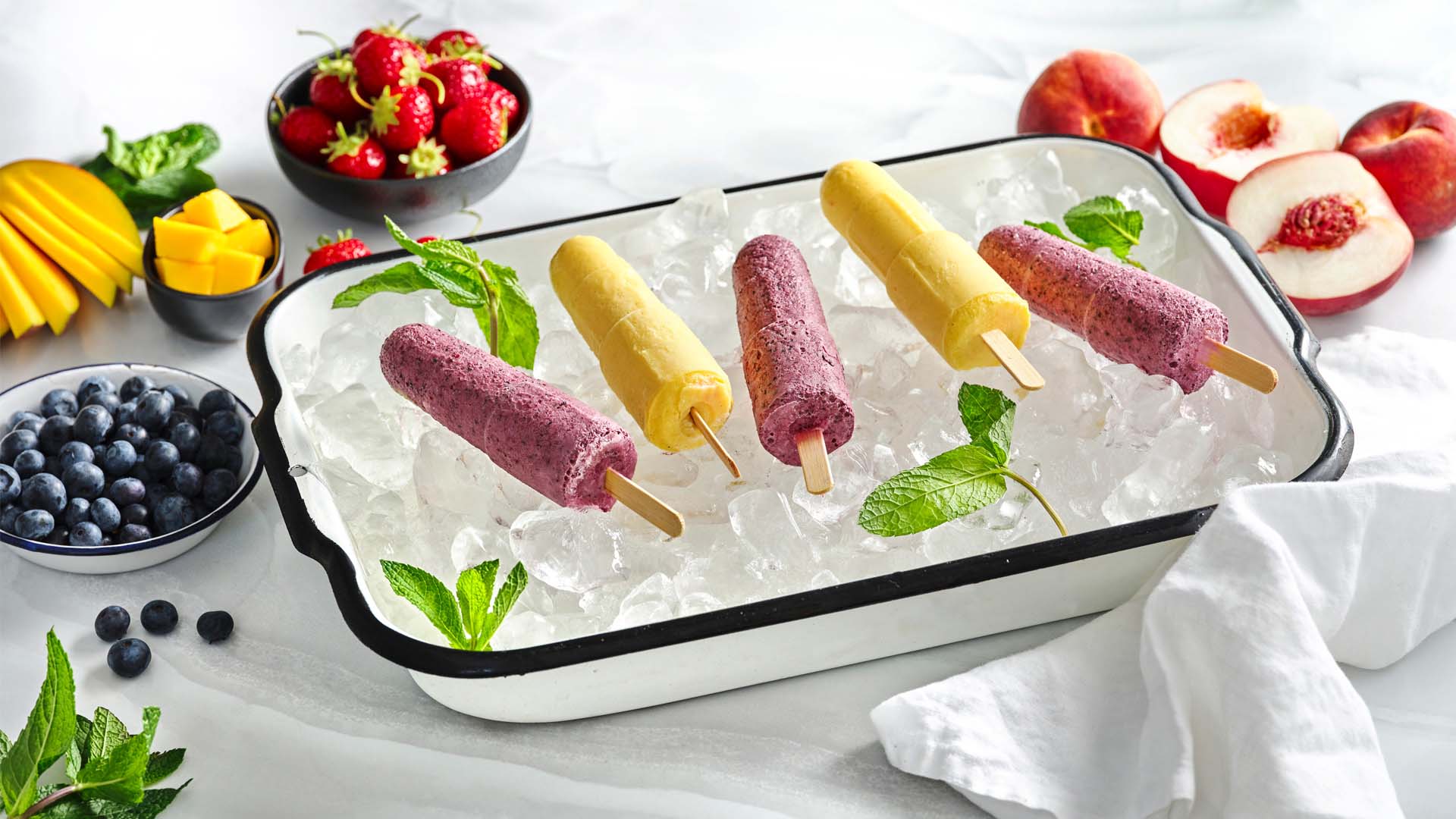 Side shot of yellow and purple fruit lassi popsicles on ice in a white rectangular dish.