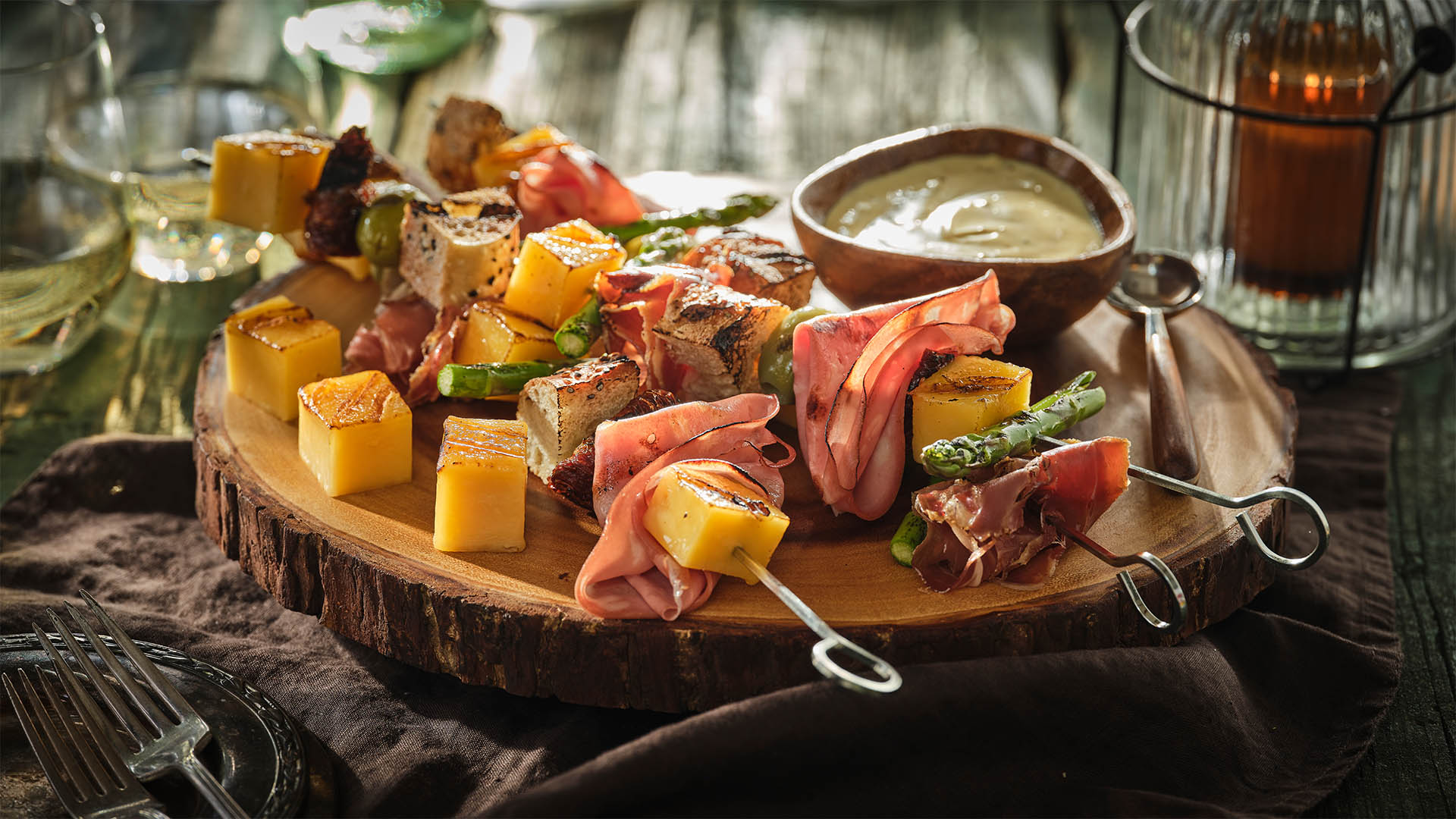 Metal Skewers on a round living edge wood board with sliced meats, cubed cheese, and bread pieces. 
